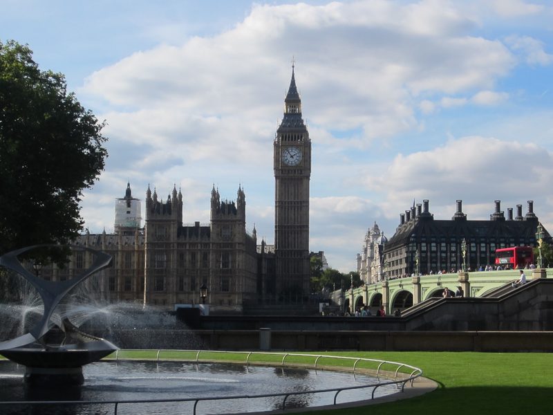 The clock tower and Gothic towers of the Palace of Westminster under a bright sky with white clouds, with Westminster Bridge and a red bus at right and a fountain on a lawn in the foreground