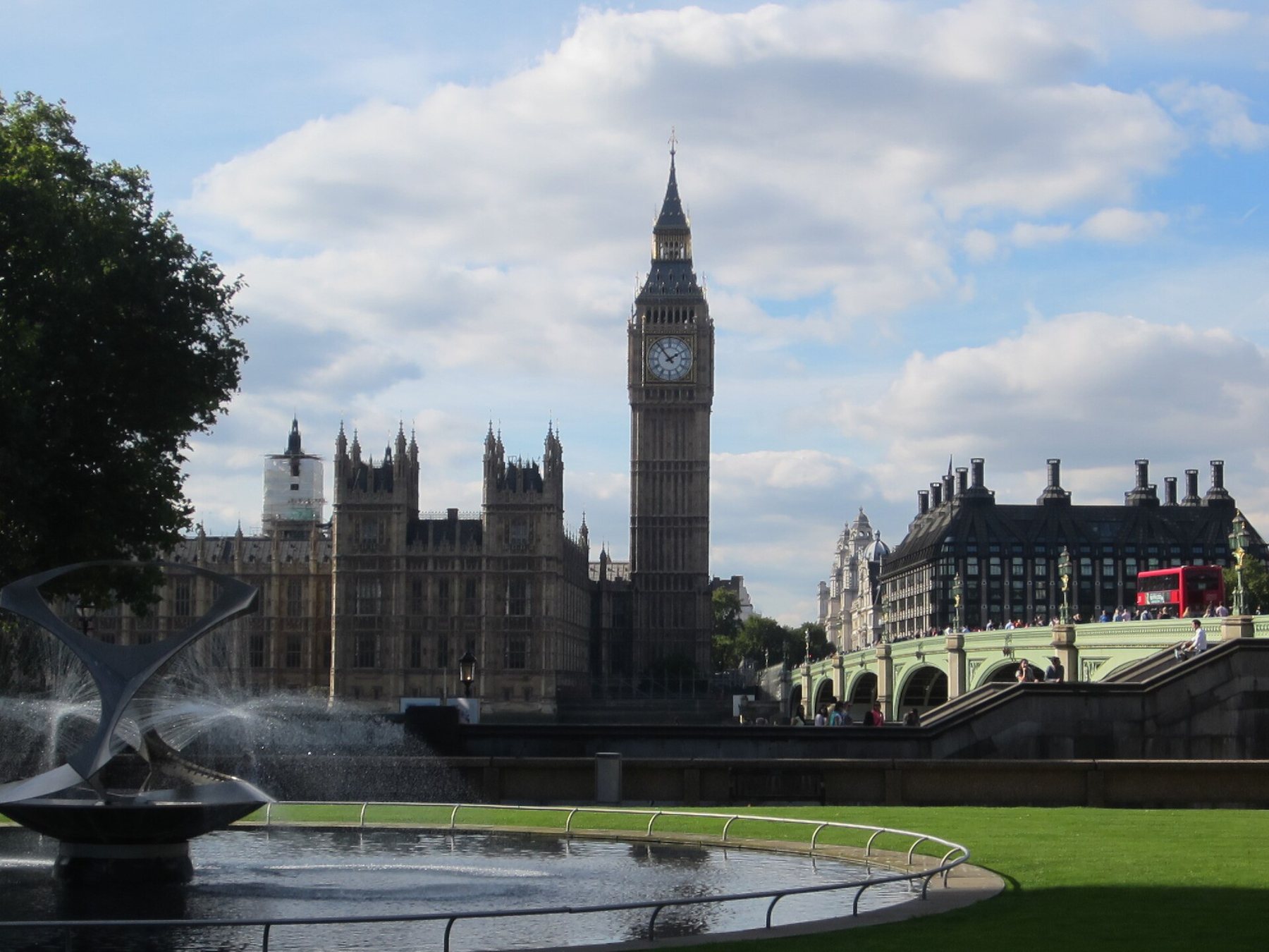 The clock tower and Gothic towers of the Palace of Westminster under a bright sky with white clouds, with Westminster Bridge and a red bus at right and a fountain on a lawn in the foreground