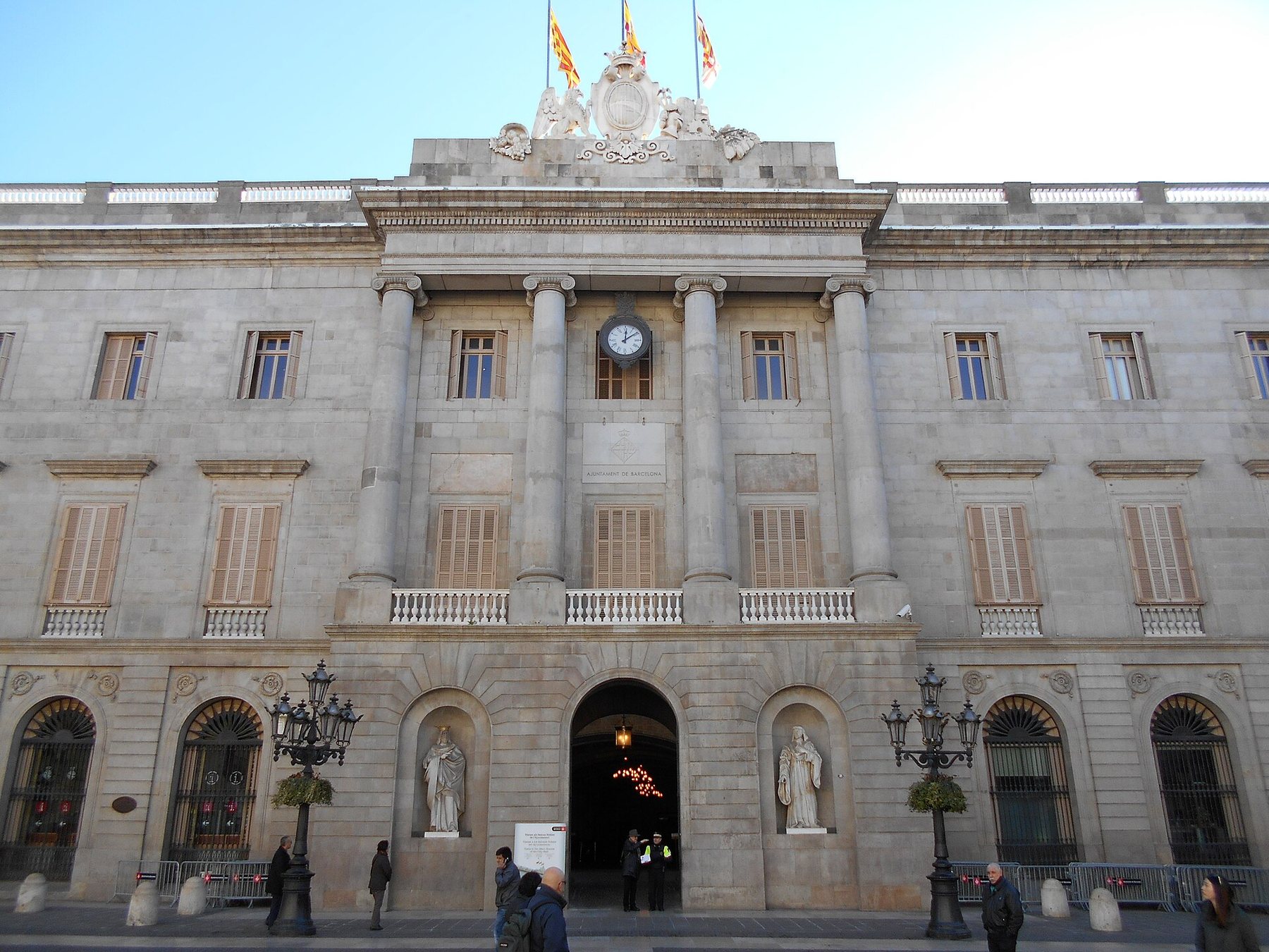 The stone facade of Barcelona City Hall in daylight: four tall columns and a clock above an arched doorway, a statue in a niche on each side of the door, two iron lamp posts in front and three flags on the roof.