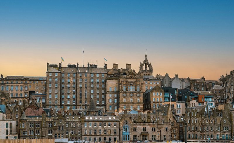 Tall stone buildings of Edinburgh's Old Town rising in tiers up a slope, with flags flying from the rooftops and an open crown spire behind them, under a clear sky fading from blue to pale orange