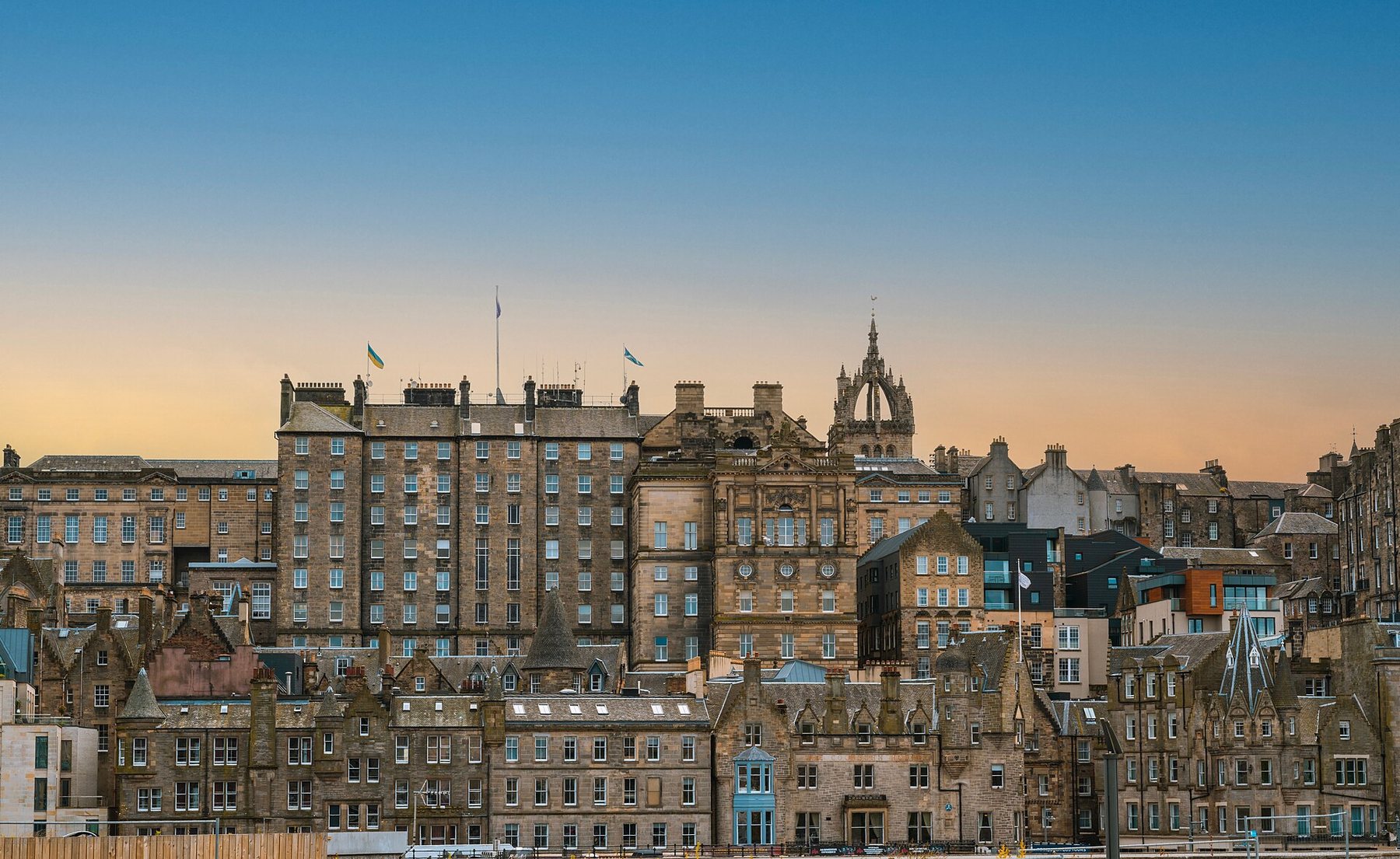 Tall stone buildings of Edinburgh's Old Town rising in tiers up a slope, with flags flying from the rooftops and an open crown spire behind them, under a clear sky fading from blue to pale orange