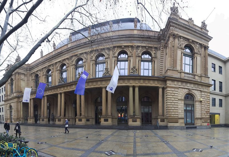 The curved sandstone front of the Frankfurt Stock Exchange building, with arched windows, columns and rooftop statues, flags hanging over the entrance and a wet paved square in front on an overcast day