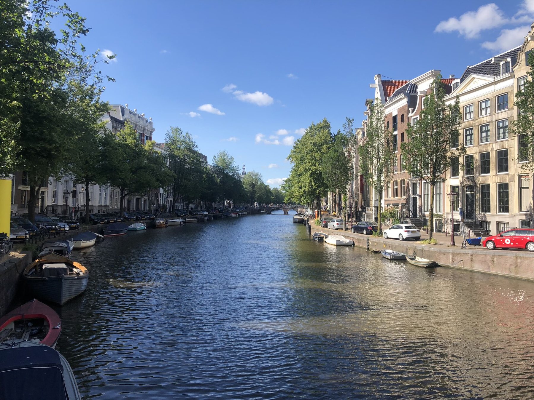 A wide Amsterdam canal on a sunny day, lined with trees, moored boats and tall brick and stone canal houses, with an arched bridge in the distance