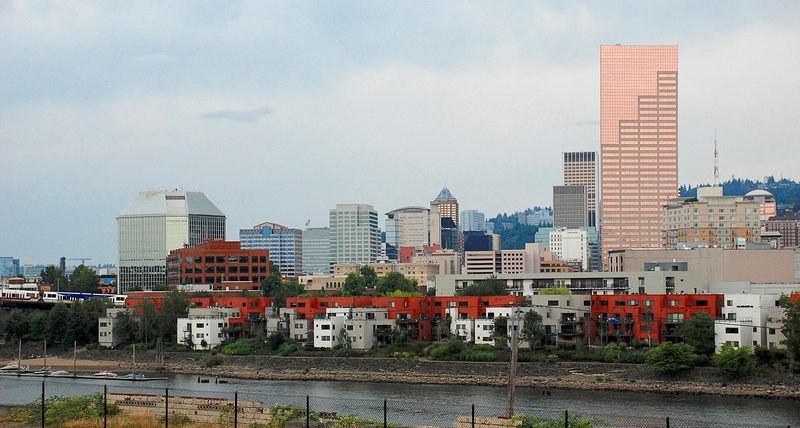 Downtown Portland's office towers, one of them a tall pink glass tower, rising behind red and white riverside apartment blocks on the Willamette River under a grey sky