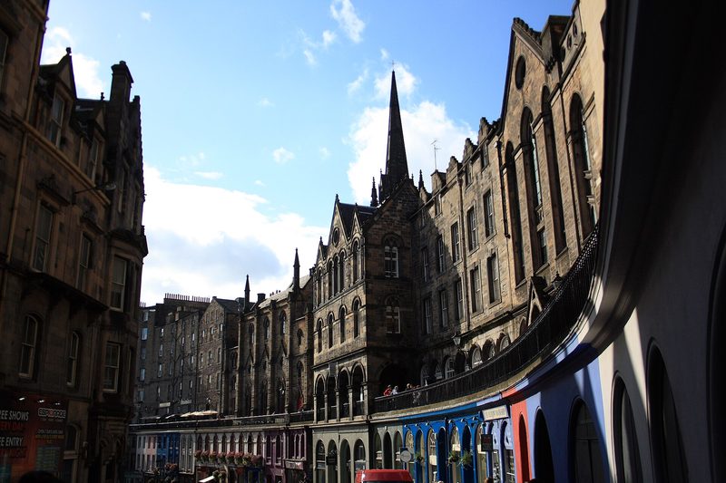 A curving row of tall sandstone buildings with a church spire behind them, above arched shopfronts painted blue and red, under a bright blue sky on Victoria Street in Edinburgh.