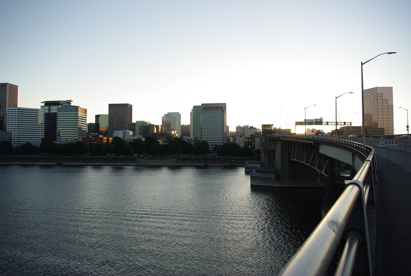 Office towers of downtown Portland line the far bank of the Willamette River in evening light, with the Morrison Bridge and its steel railing curving in from the right.