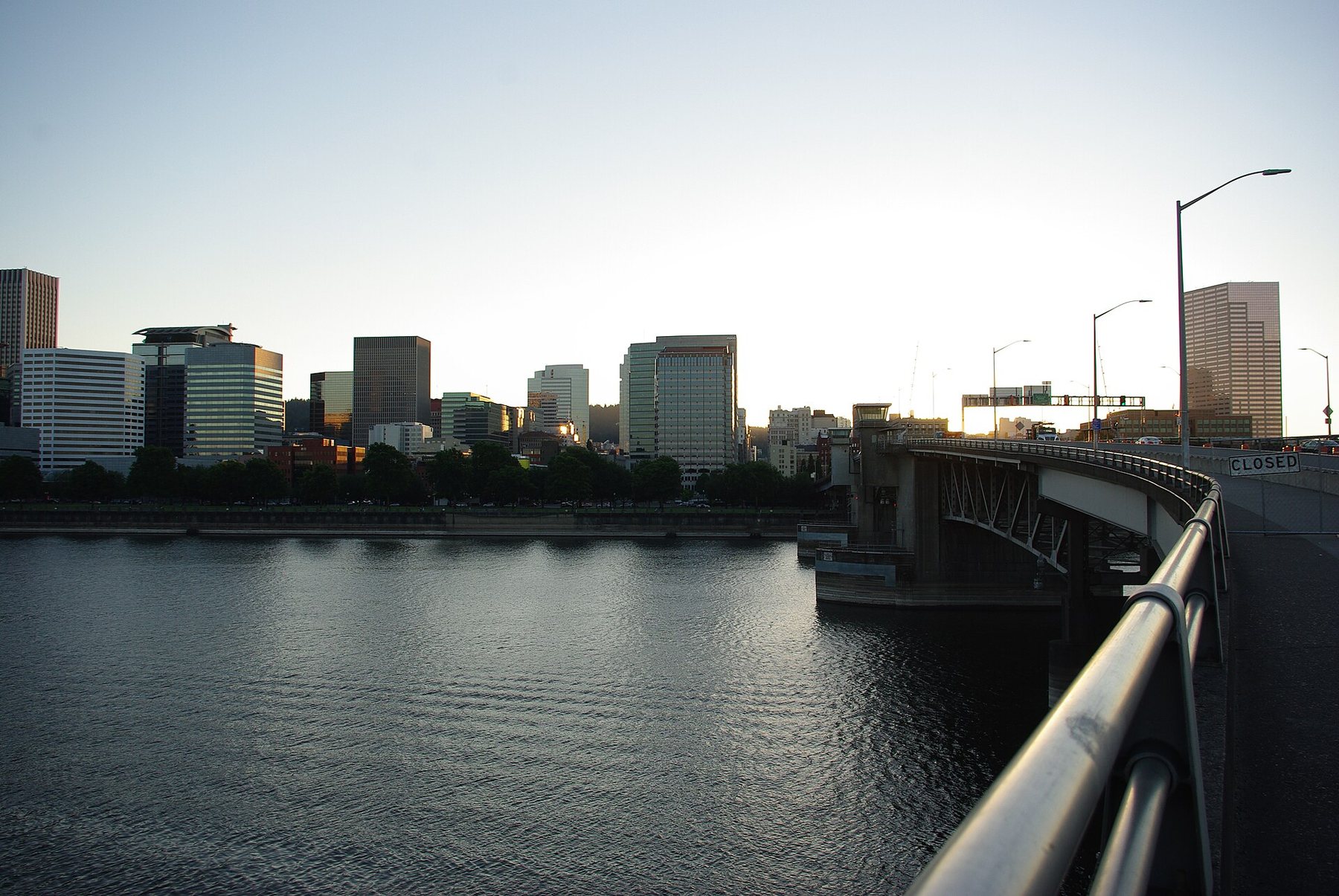 Office towers of downtown Portland line the far bank of the Willamette River in evening light, with the Morrison Bridge and its steel railing curving in from the right.