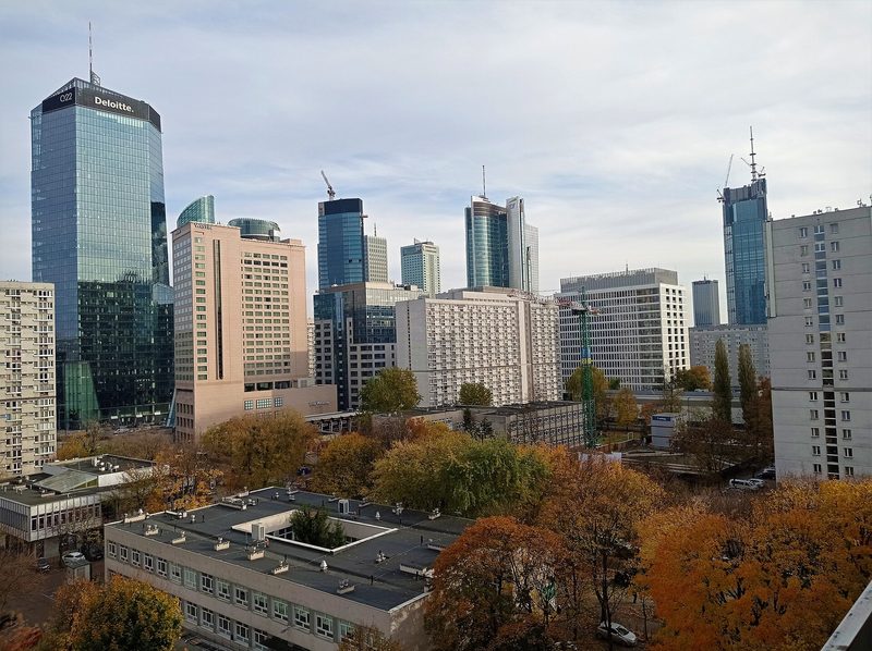 Glass office towers and pale concrete apartment blocks in central Warsaw rise above trees in autumn colour under a light cloudy sky.