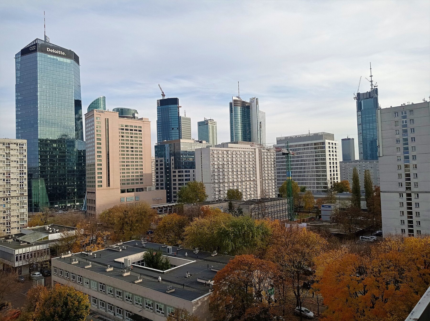 Glass office towers and pale concrete apartment blocks in central Warsaw rise above trees in autumn colour under a light cloudy sky.