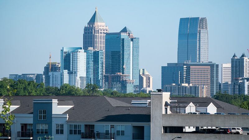 Glass and stone office towers of Midtown Atlanta rise under a clear blue sky behind a line of trees, with the dark roofs of a low apartment complex and a parking deck in the foreground.