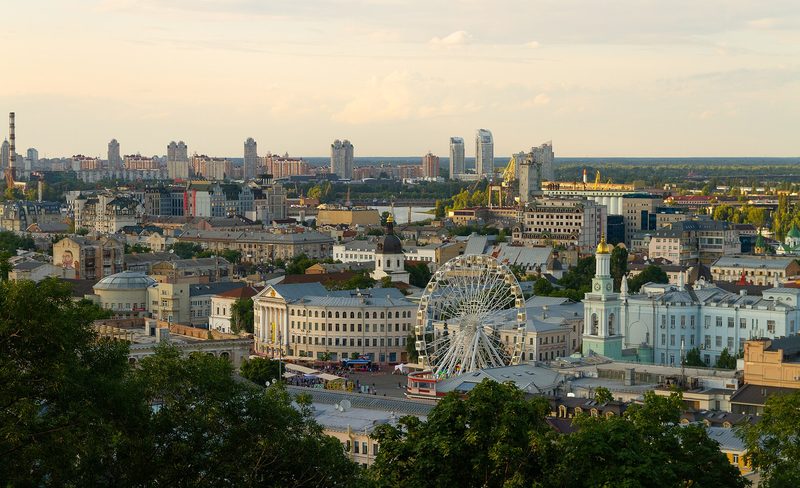 A wide view over the low rooftops of central Kyiv in evening light, with a white Ferris wheel on Kontraktova Square in the middle, a pale blue bell tower to its right and residential towers on the horizon.