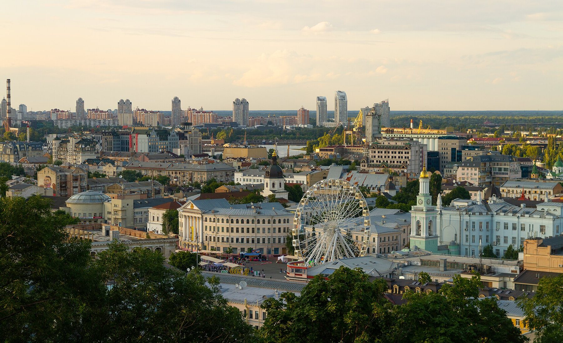 A wide view over the low rooftops of central Kyiv in evening light, with a white Ferris wheel on Kontraktova Square in the middle, a pale blue bell tower to its right and residential towers on the horizon.