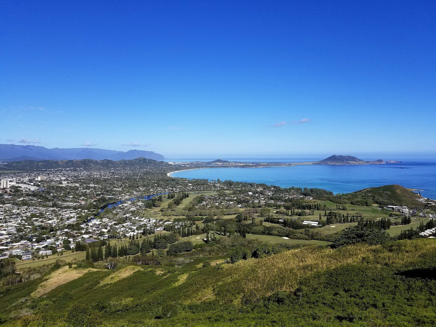 View from a green hillside over the town of Kailua on Oahu: low houses among trees, a golf course, the curve of Kailua Bay and a peninsula under a clear blue sky.
