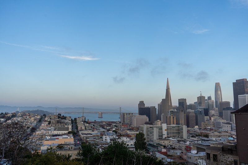 View over low rooftops to the San Francisco skyline at dusk, with the pointed Transamerica Pyramid, taller towers to its right and the Bay Bridge across the water on the left.