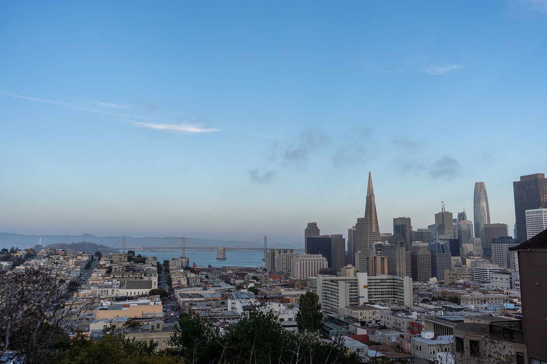 View over low rooftops to the San Francisco skyline at dusk, with the pointed Transamerica Pyramid, taller towers to its right and the Bay Bridge across the water on the left.