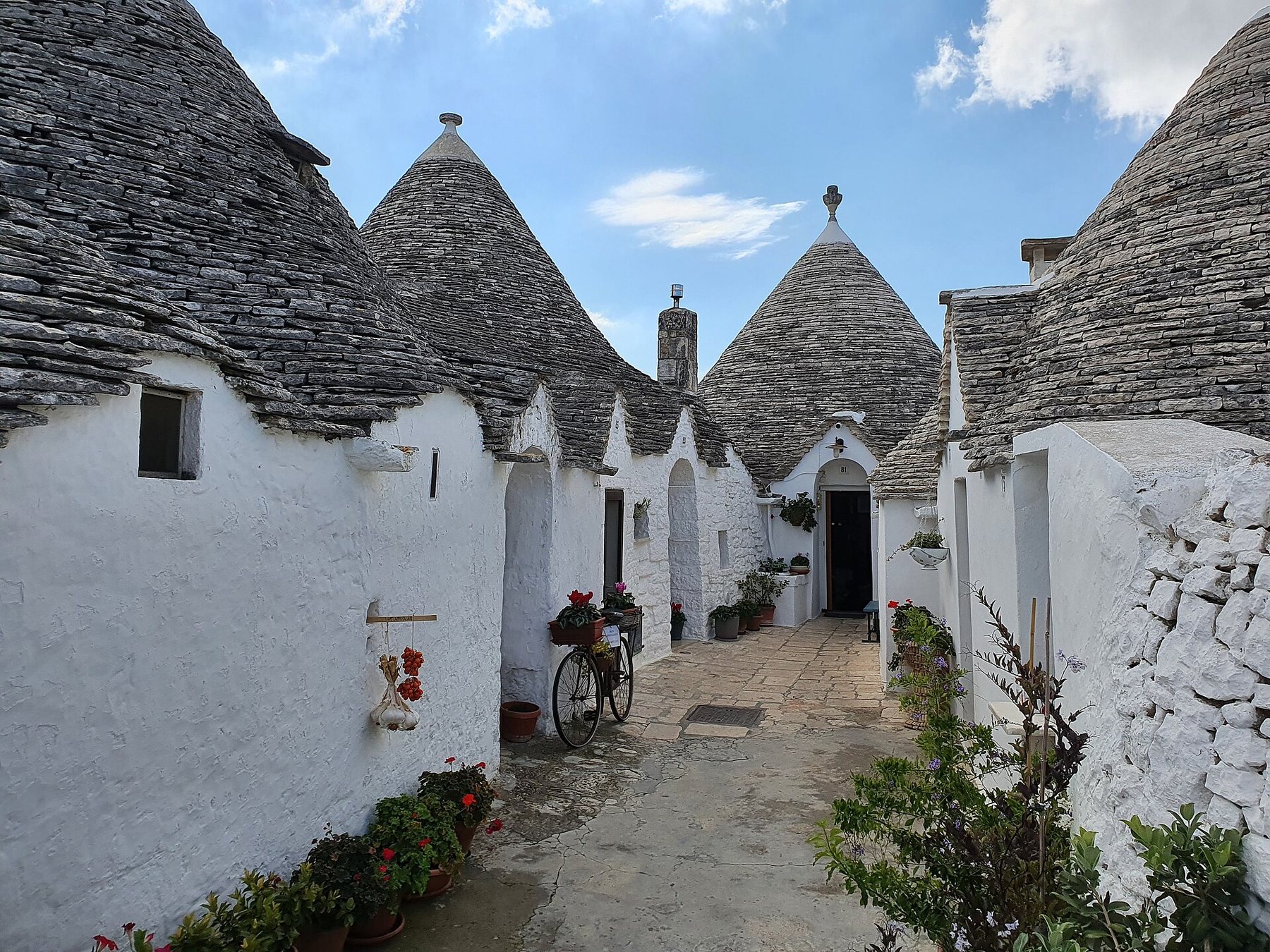 A narrow paved lane between whitewashed trulli, small stone houses with conical grey roofs, with flower pots and a parked bicycle under a blue sky.
