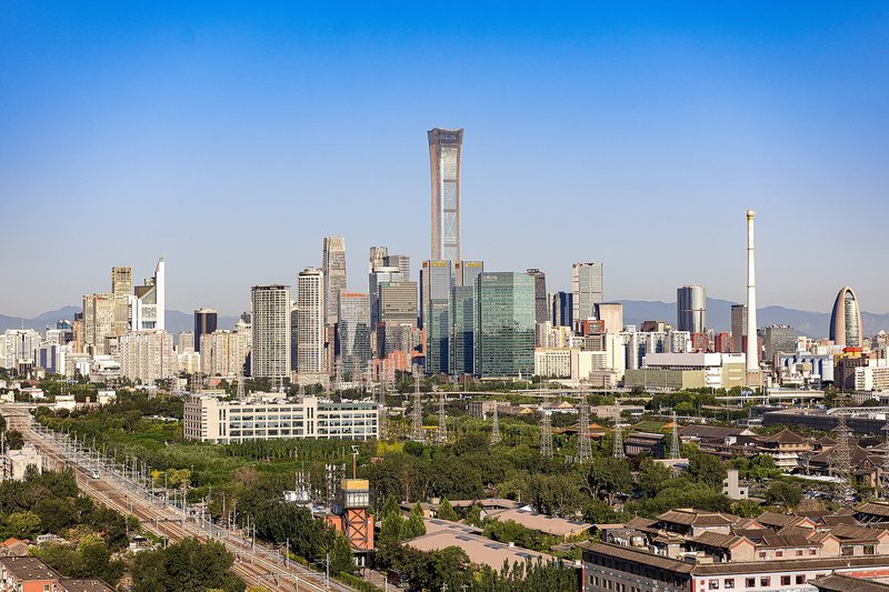 A cluster of glass office towers in Beijing's central business district under a clear blue sky, with railway lines and low tiled roofs in the foreground.