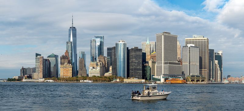 The towers of Lower Manhattan seen across the water from Governors Island under a cloudy sky, with a small white motorboat in the foreground.