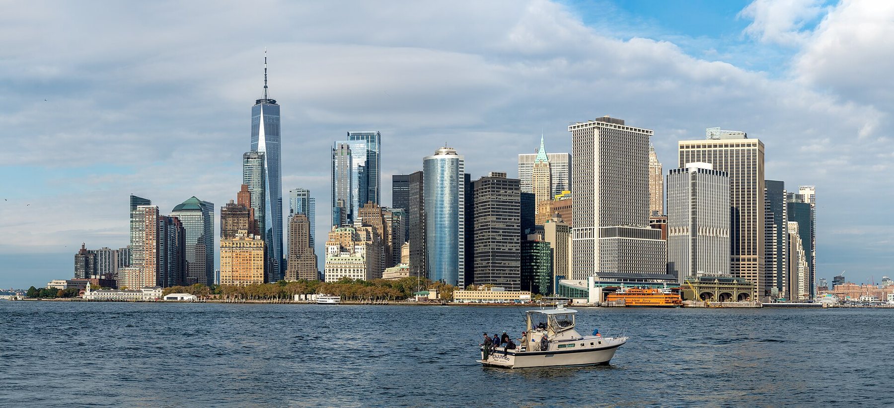 The towers of Lower Manhattan seen across the water from Governors Island under a cloudy sky, with a small white motorboat in the foreground.