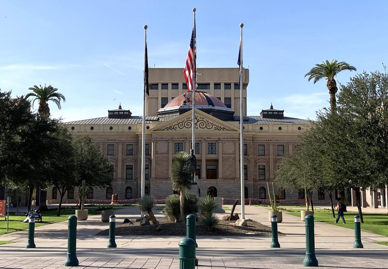 The stone front of the Arizona State Capitol in Phoenix under a clear sky, with its copper dome, three flagpoles, palm trees and the taller executive tower rising behind.
