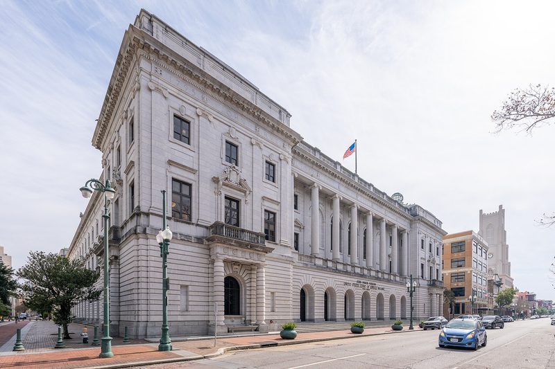 A large pale stone courthouse with a long row of tall columns and arched ground-floor openings stands on a street corner, an American flag flying from its roof and cars parked along the kerb.
