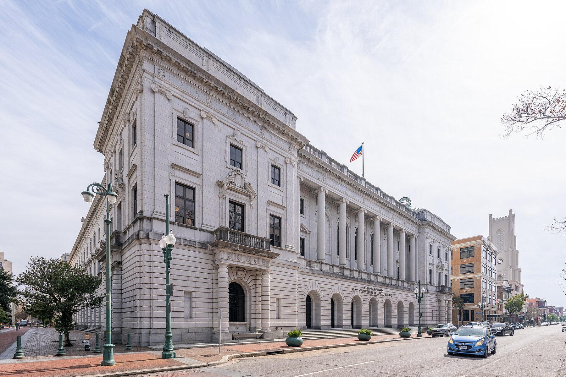 A large pale stone courthouse with a long row of tall columns and arched ground-floor openings stands on a street corner, an American flag flying from its roof and cars parked along the kerb.