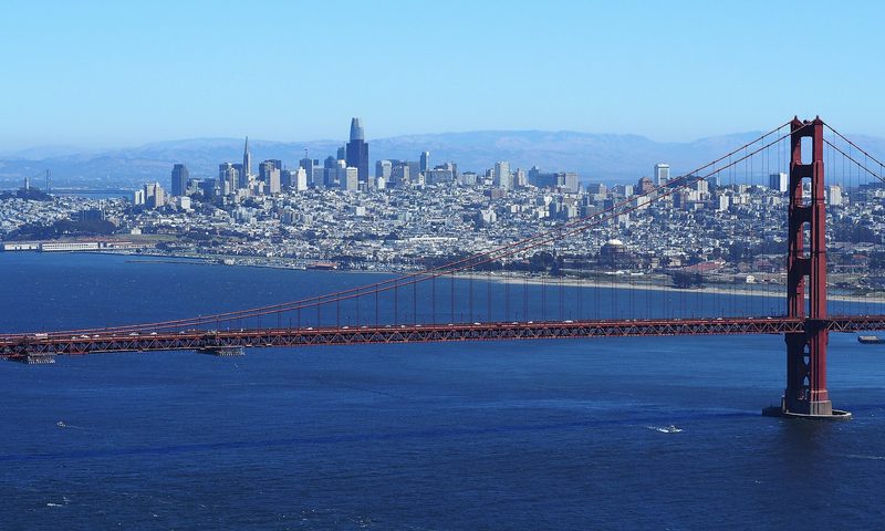 The towers of downtown San Francisco rise beyond the red span and south tower of the Golden Gate Bridge, seen across blue water under a clear sky.