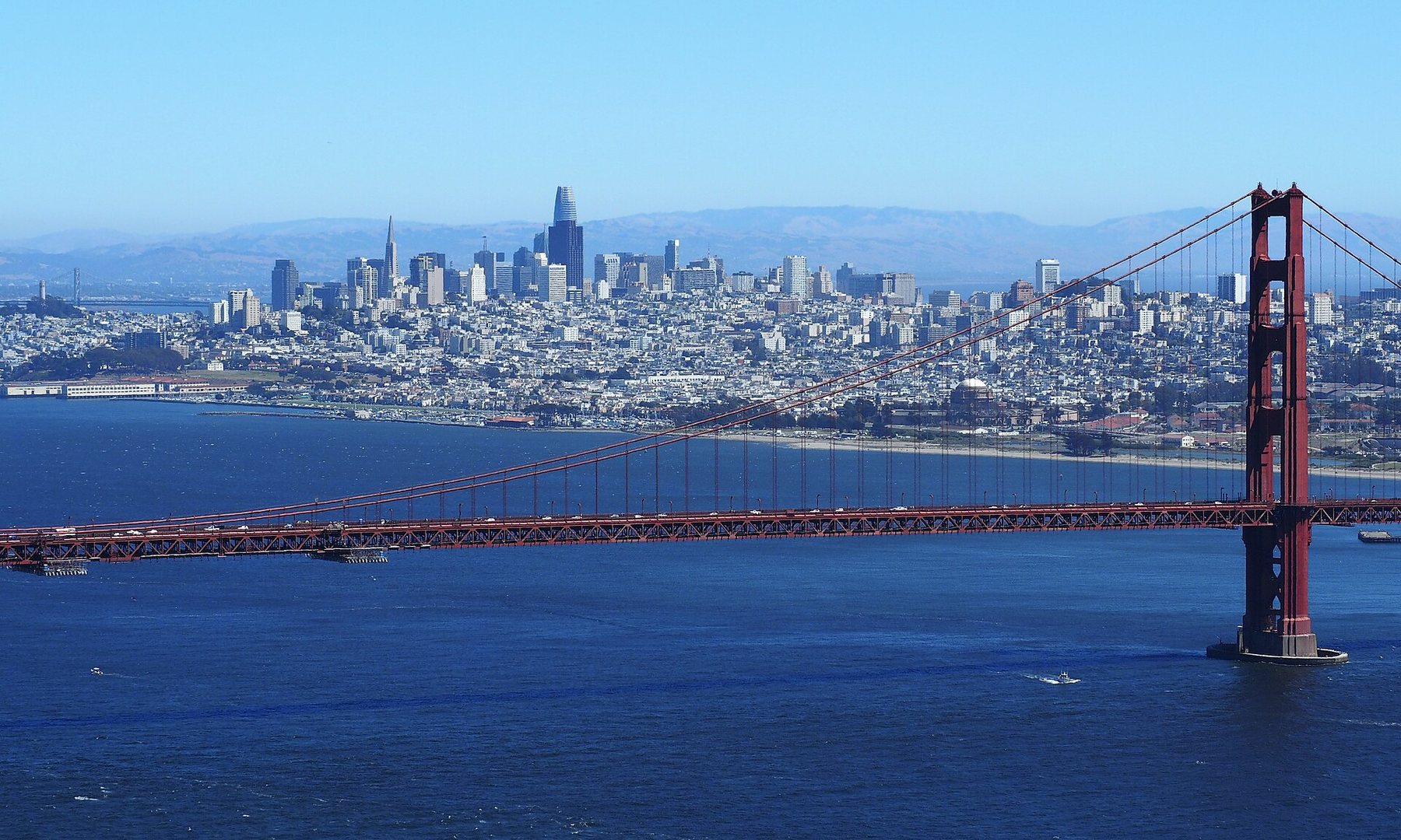 The towers of downtown San Francisco rise beyond the red span and south tower of the Golden Gate Bridge, seen across blue water under a clear sky.