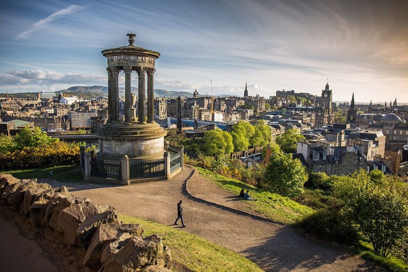 A view over central Edinburgh in low evening sun: a round, columned stone monument on a grassy hill in the foreground, with stone rooftops, a clock tower and the castle on the skyline.