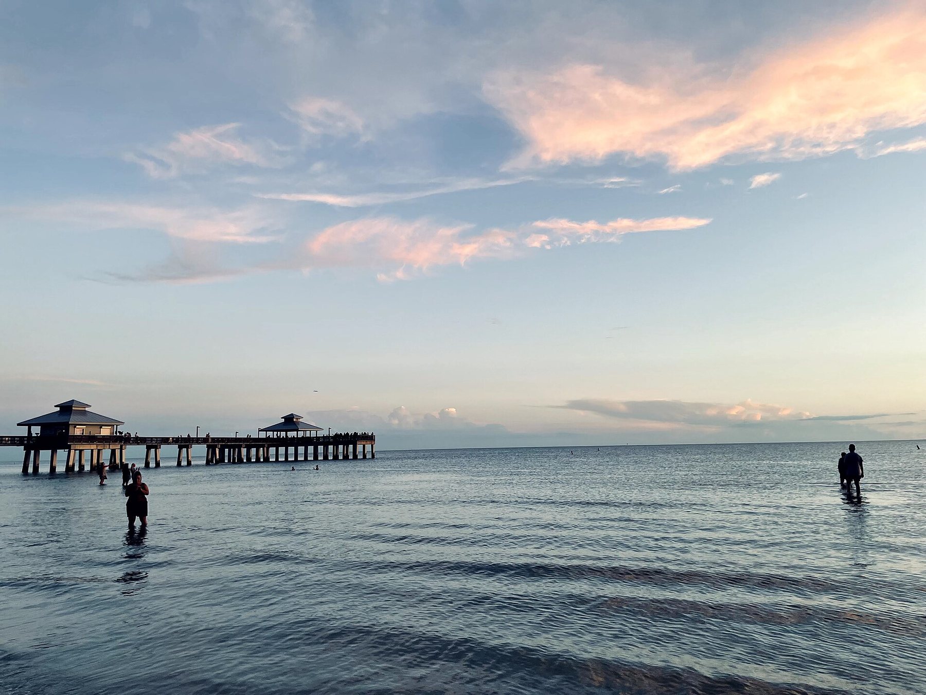 A long fishing pier with two roofed pavilions reaches into calm Gulf water at dusk, with a few people wading in the shallows under pink-edged clouds.