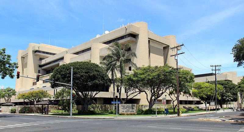 A large tan concrete building with rounded corner towers stands behind palm and shade trees at a street corner under a blue sky.