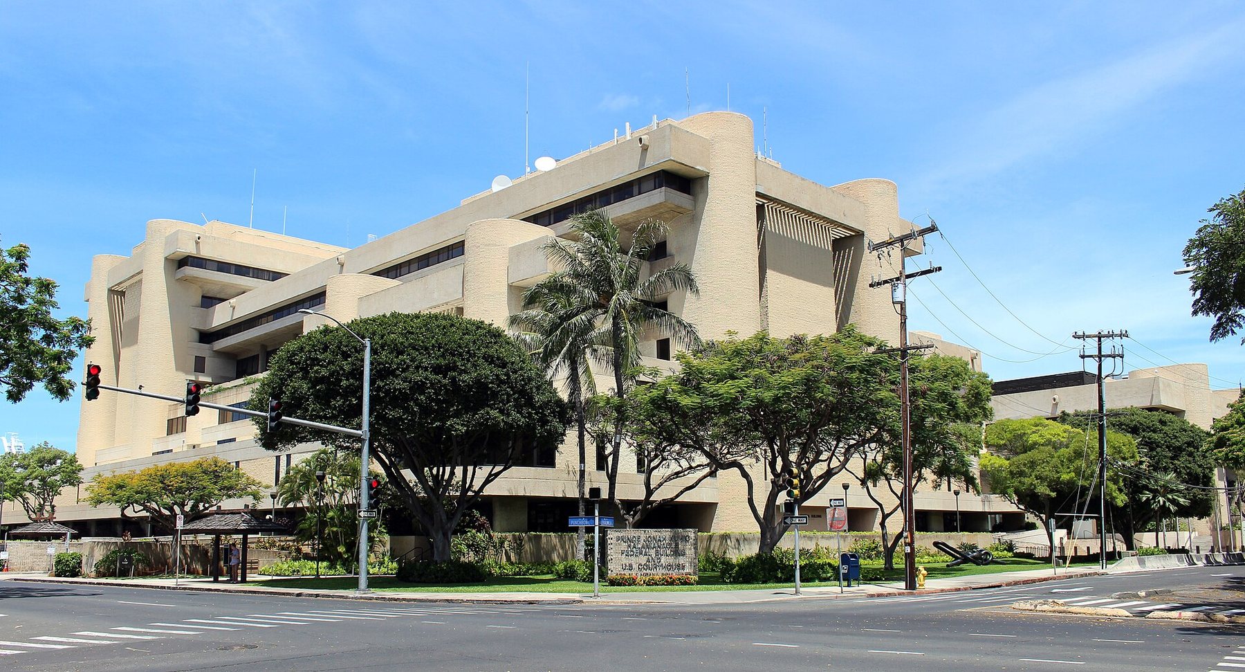 A large tan concrete building with rounded corner towers stands behind palm and shade trees at a street corner under a blue sky.