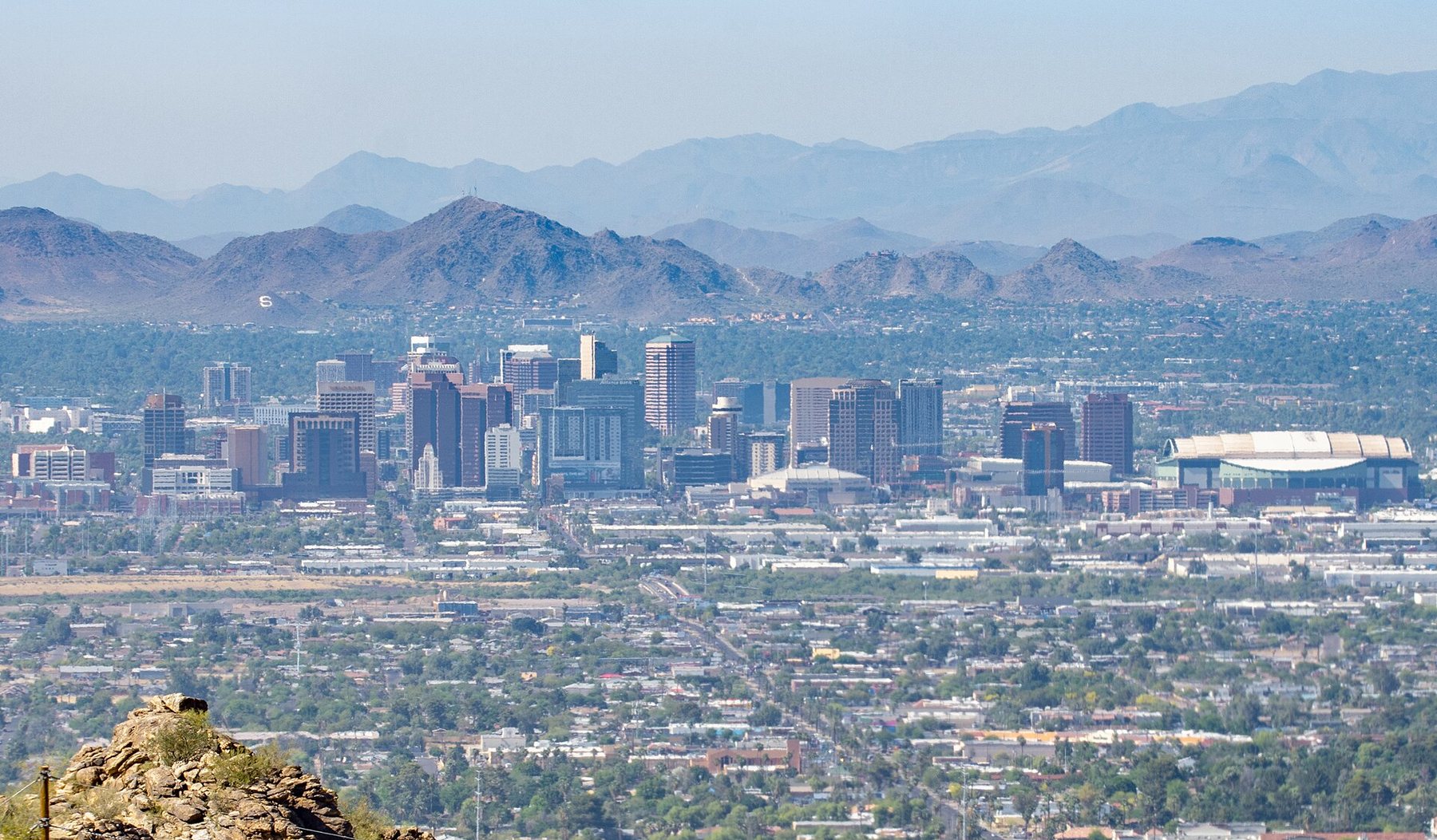 A wide view across low-rise neighborhoods to the downtown Phoenix skyline, with desert mountain ranges behind it in a light haze.