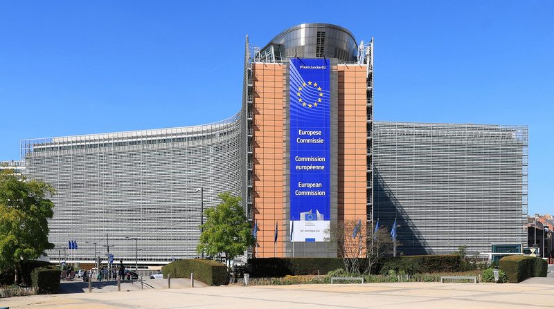 A wide curved office building of glass and steel under a clear blue sky, with a tall blue banner down its centre showing the ring of gold stars and the words European Commission in three languages.