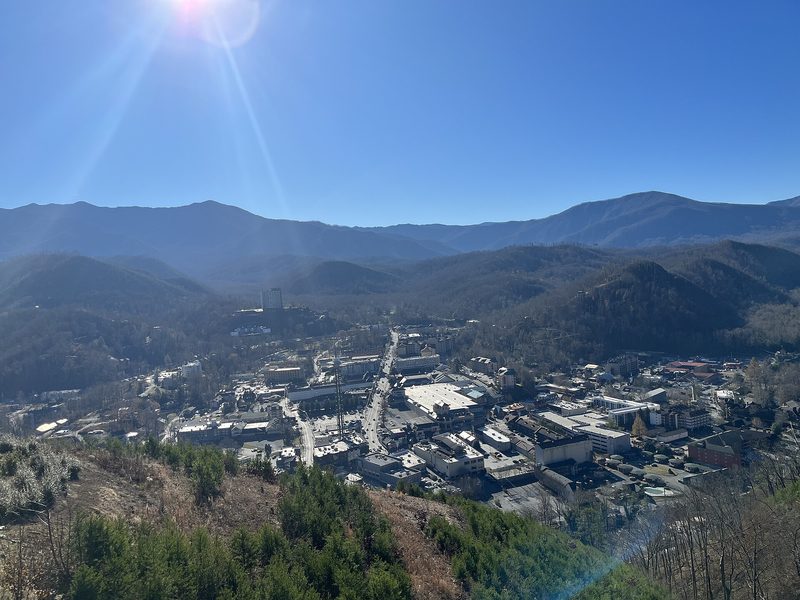 The town of Gatlinburg, Tennessee, seen from a hillside above it: a main street and low buildings in a valley between wooded mountain ridges under a clear blue sky.