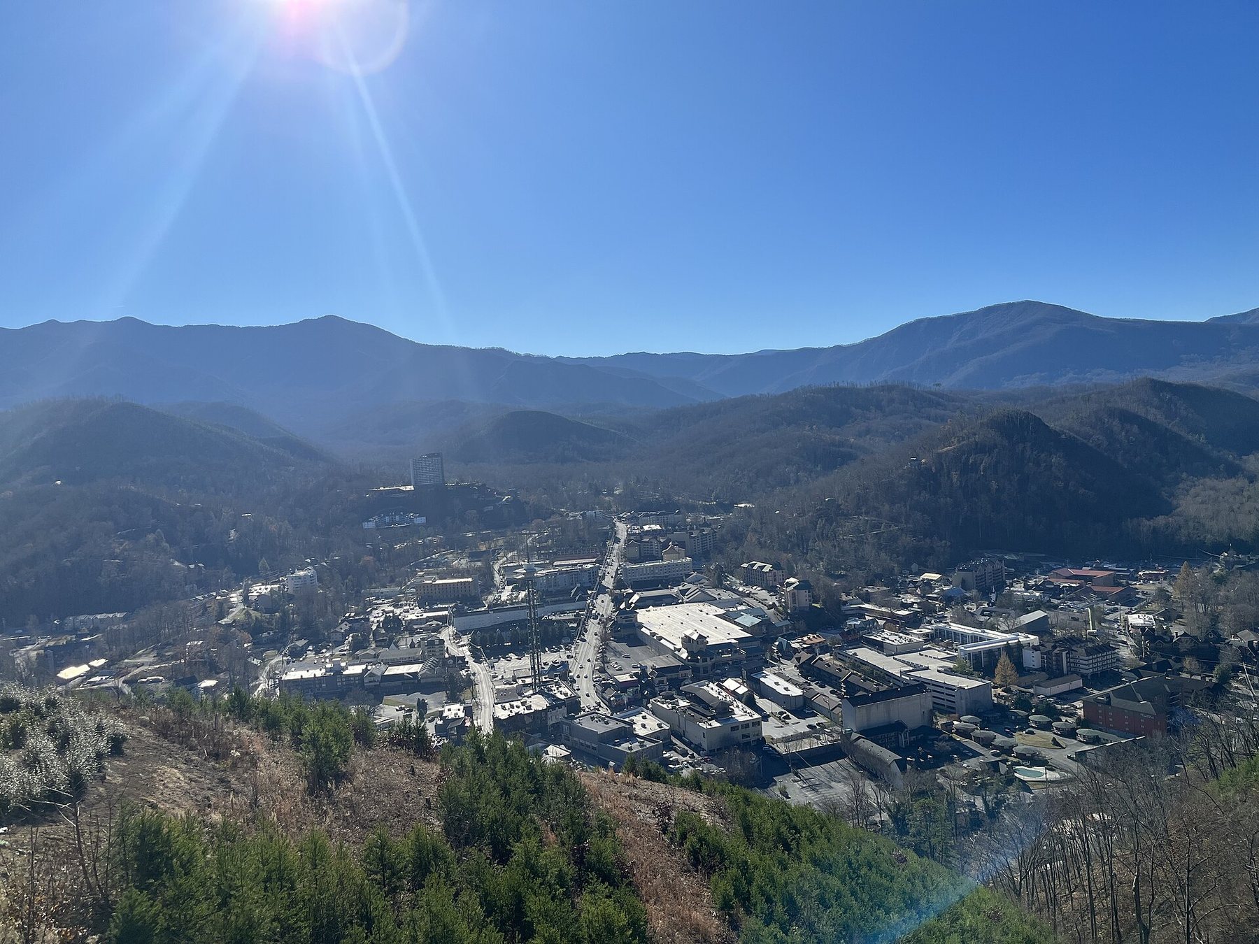 The town of Gatlinburg, Tennessee, seen from a hillside above it: a main street and low buildings in a valley between wooded mountain ridges under a clear blue sky.