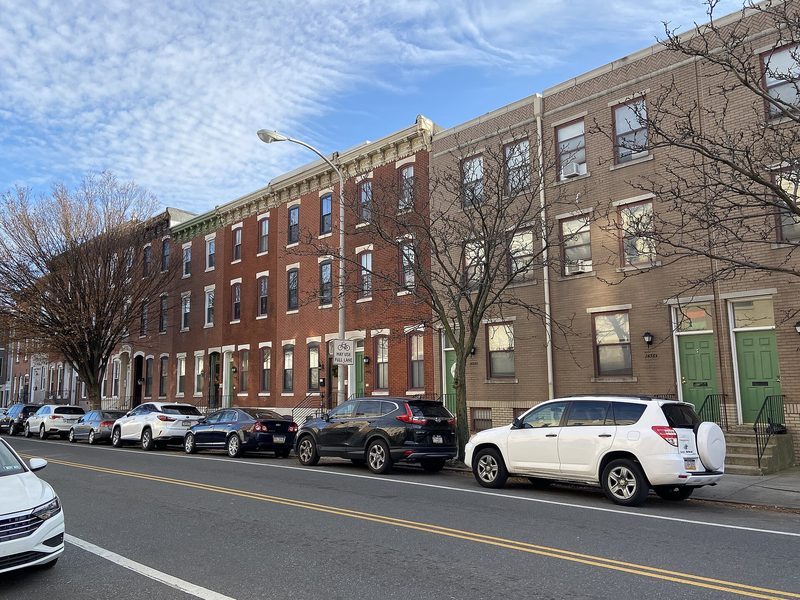Three-storey brick row homes line a Philadelphia street, with parked cars at the kerb and bare winter trees under a blue sky streaked with thin cloud.