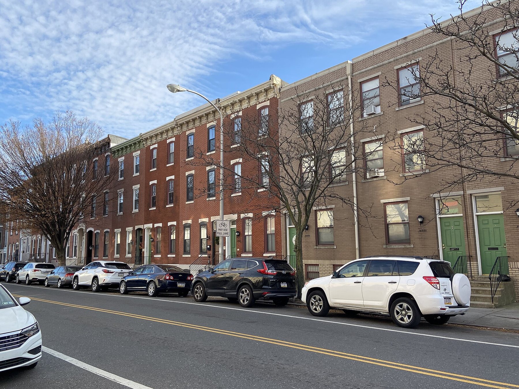 Three-storey brick row homes line a Philadelphia street, with parked cars at the kerb and bare winter trees under a blue sky streaked with thin cloud.