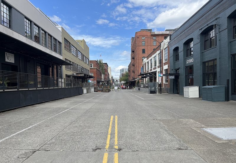 An empty stretch of NW 13th Avenue in Portland's Pearl District under a blue sky, with converted brick warehouses and raised loading docks on both sides of the street.
