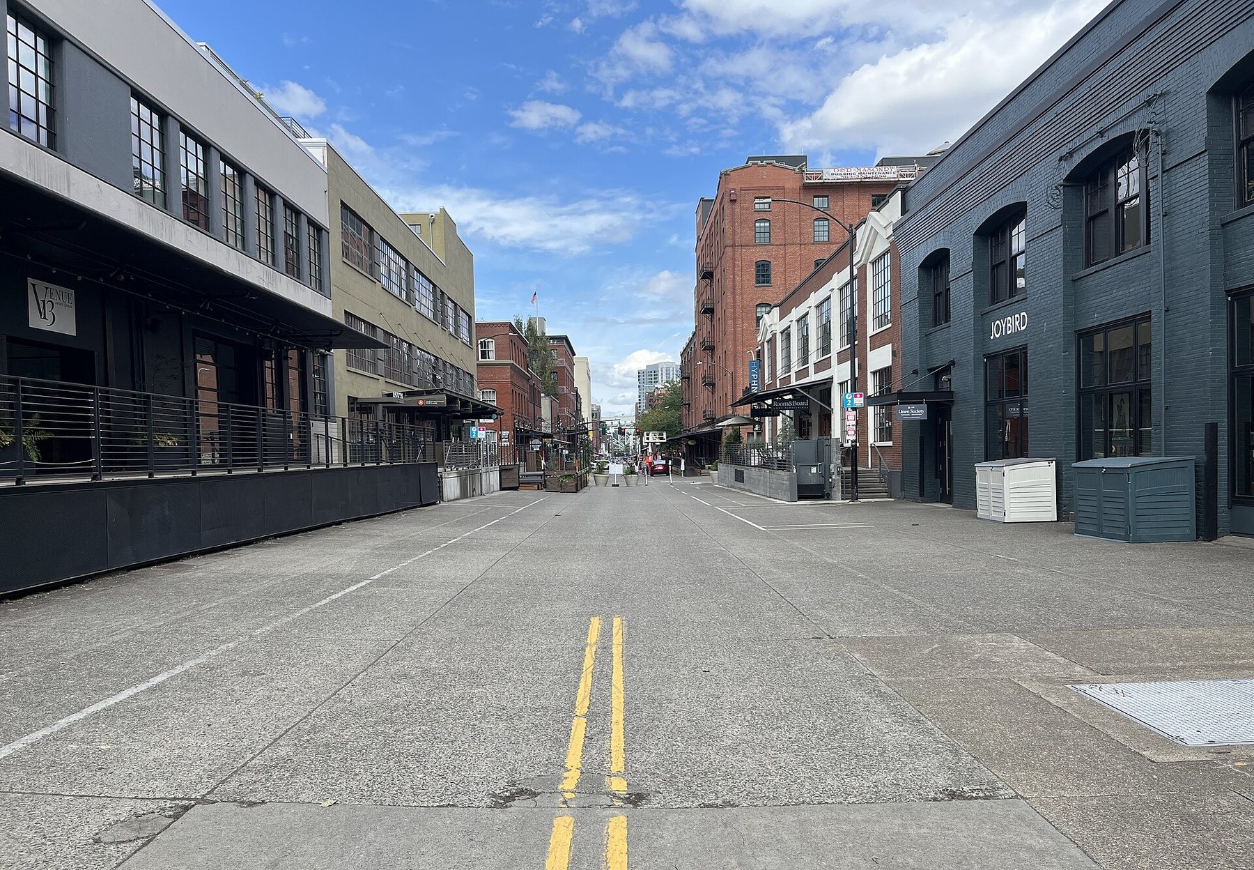 An empty stretch of NW 13th Avenue in Portland's Pearl District under a blue sky, with converted brick warehouses and raised loading docks on both sides of the street.