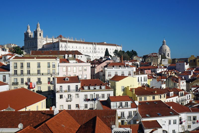 Red-tiled rooftops and white, yellow and pink apartment houses of Lisbon's Alfama district climb a hillside under a clear blue sky, with a white twin-towered church on the ridge at the left and a pale stone dome at the right.