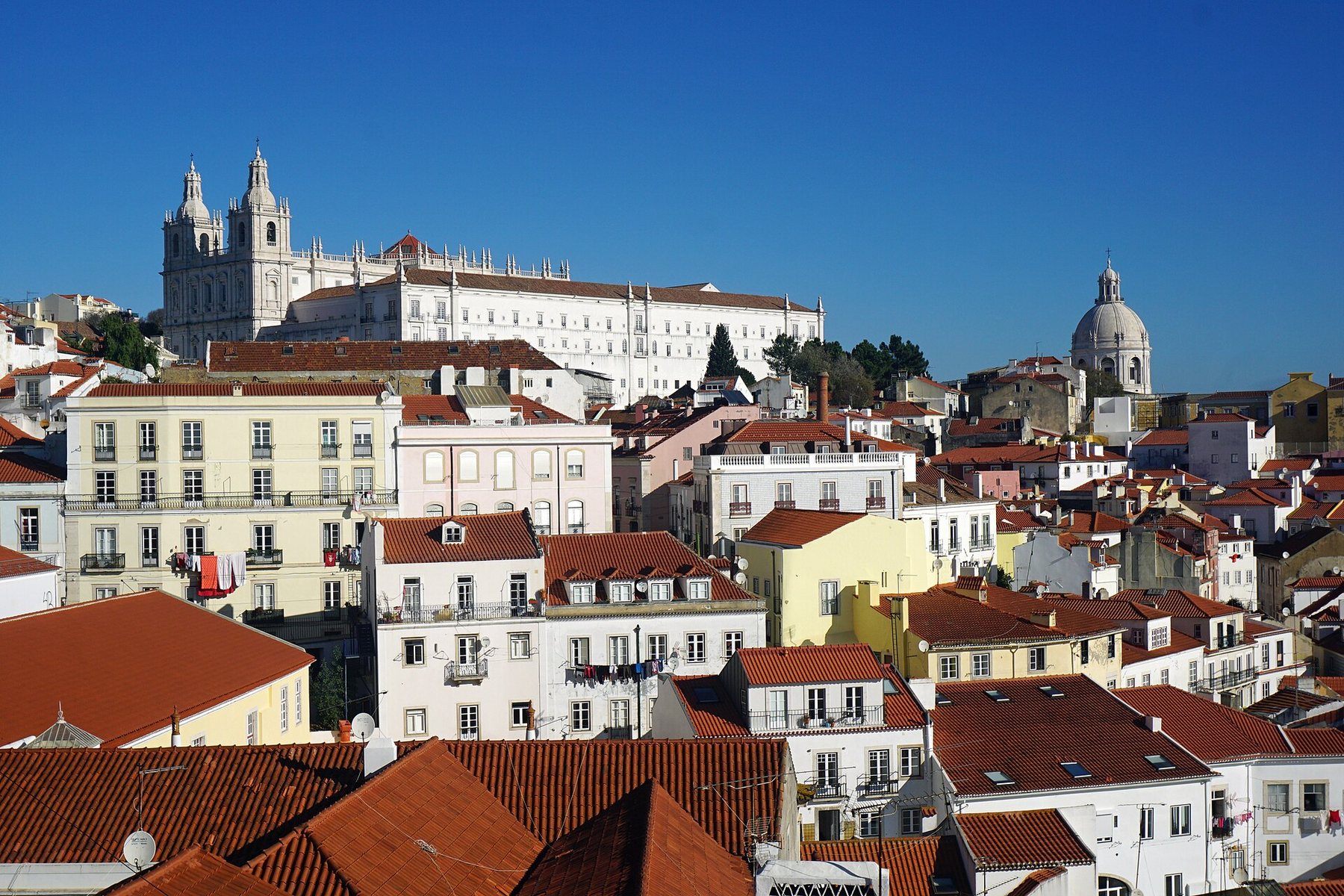 Red-tiled rooftops and white, yellow and pink apartment houses of Lisbon's Alfama district climb a hillside under a clear blue sky, with a white twin-towered church on the ridge at the left and a pale stone dome at the right.