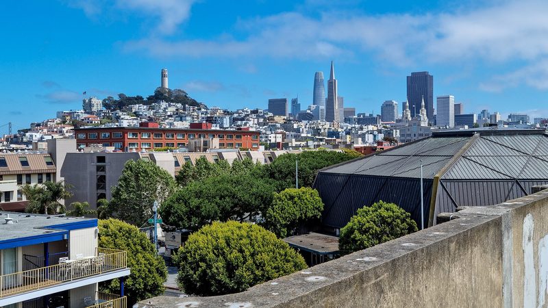 Coit Tower on its hill and the towers of downtown San Francisco, among them the Transamerica Pyramid, rise behind apartment buildings and trees under a blue sky.