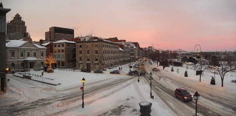 Snow-covered streets in Old Montreal at dusk: stone and brick buildings line Rue de la Commune beside Place d'Youville under a pink sky, with a Ferris wheel and a bridge in the distance.