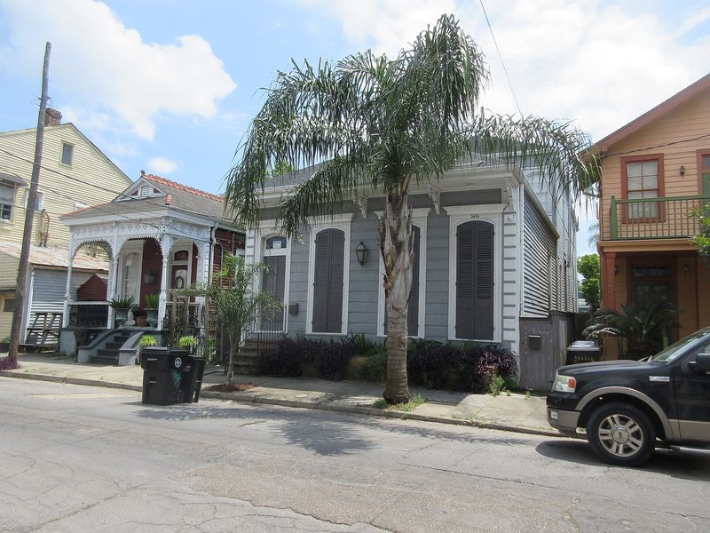 A grey wooden cottage with shuttered doors behind a palm tree, between a white-trimmed shotgun house and an orange two-storey house, on a residential street in New Orleans.