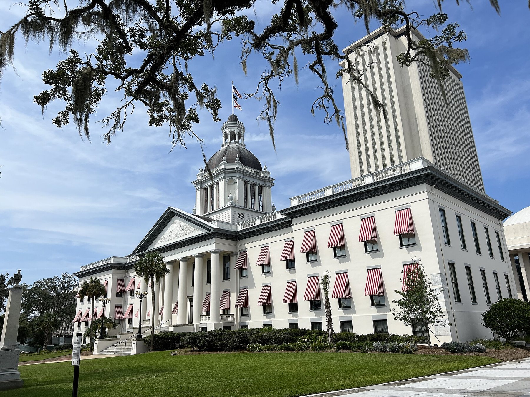 The old Florida capitol in Tallahassee, a white columned building with a dome and red-and-white striped window awnings, with the tall newer capitol tower rising behind it under a blue sky, framed by oak branches hung with Spanish moss.