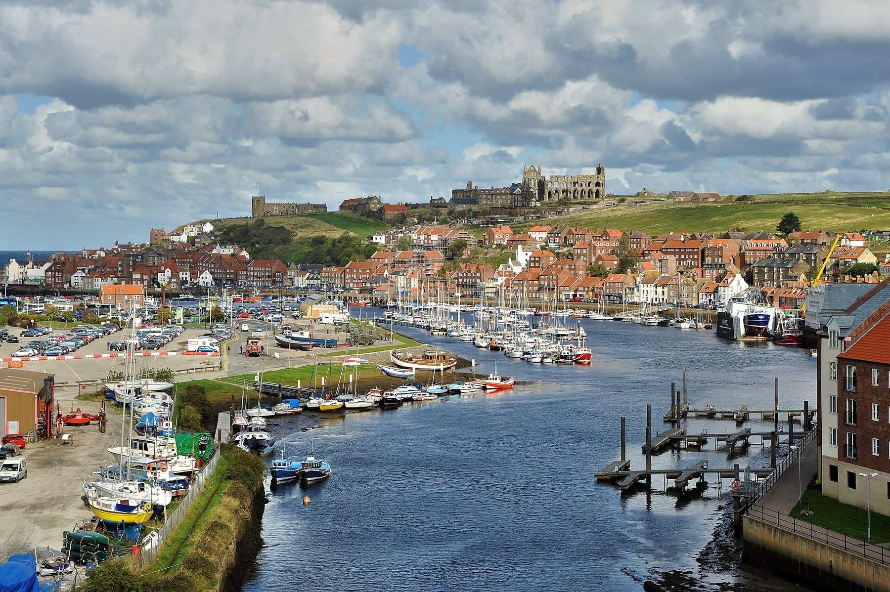 The River Esk at Whitby with rows of small boats moored along both banks, red-roofed houses climbing the far hillside and the ruins of Whitby Abbey on the skyline under broken cloud.