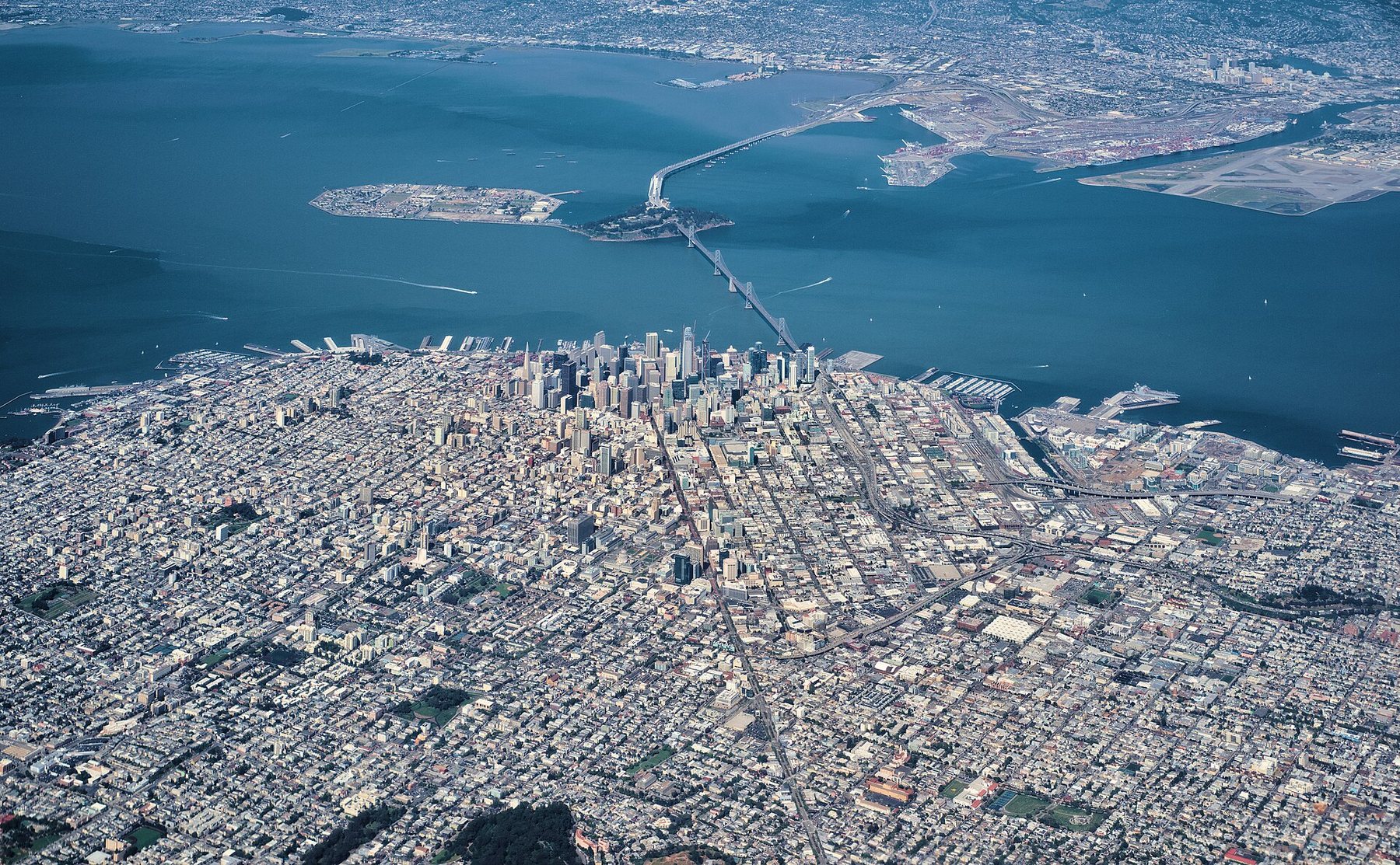 Aerial view of a dense city grid on a peninsula, with a cluster of downtown towers at the water's edge, a long bridge crossing the bay by way of a small island, and a second city on the far shore.
