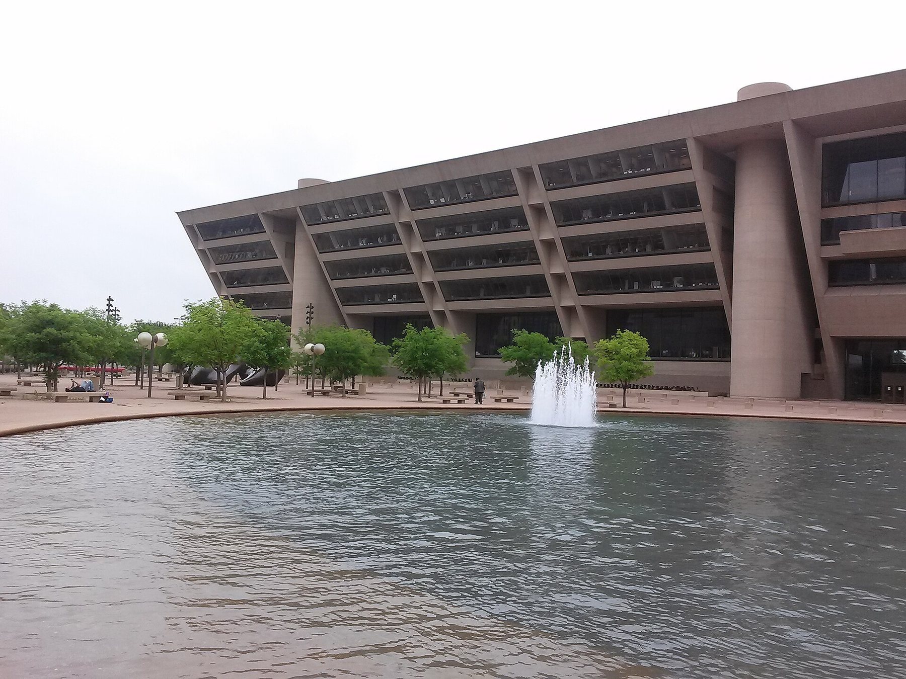 Dallas City Hall, a concrete building whose front leans out over its plaza, seen across a pool with a fountain and a row of young trees under an overcast sky.