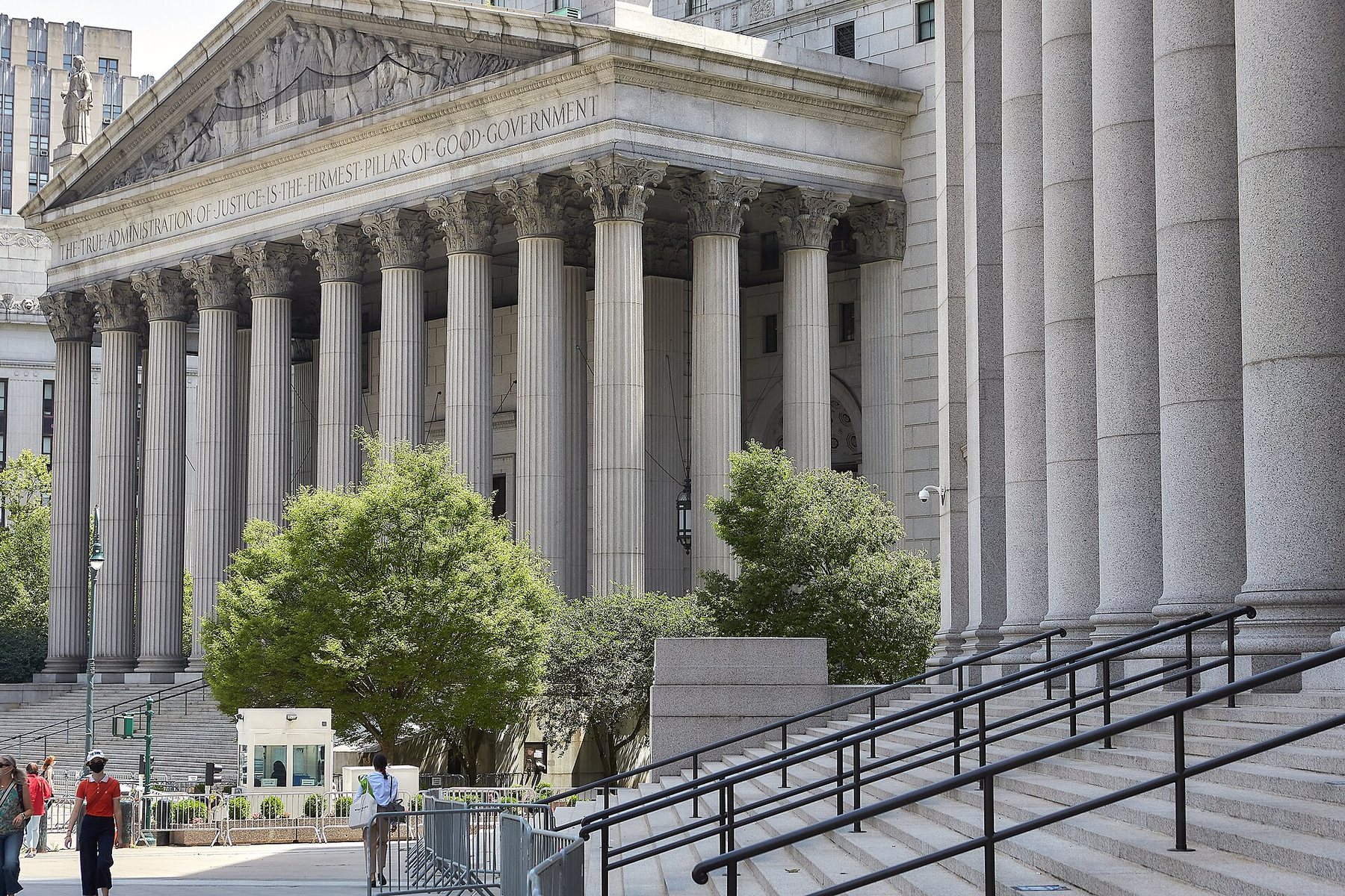 The columned front of the New York County Supreme Court in Manhattan, with an inscription under its carved pediment, seen past the steps and columns of a neighbouring building, with green trees in front.